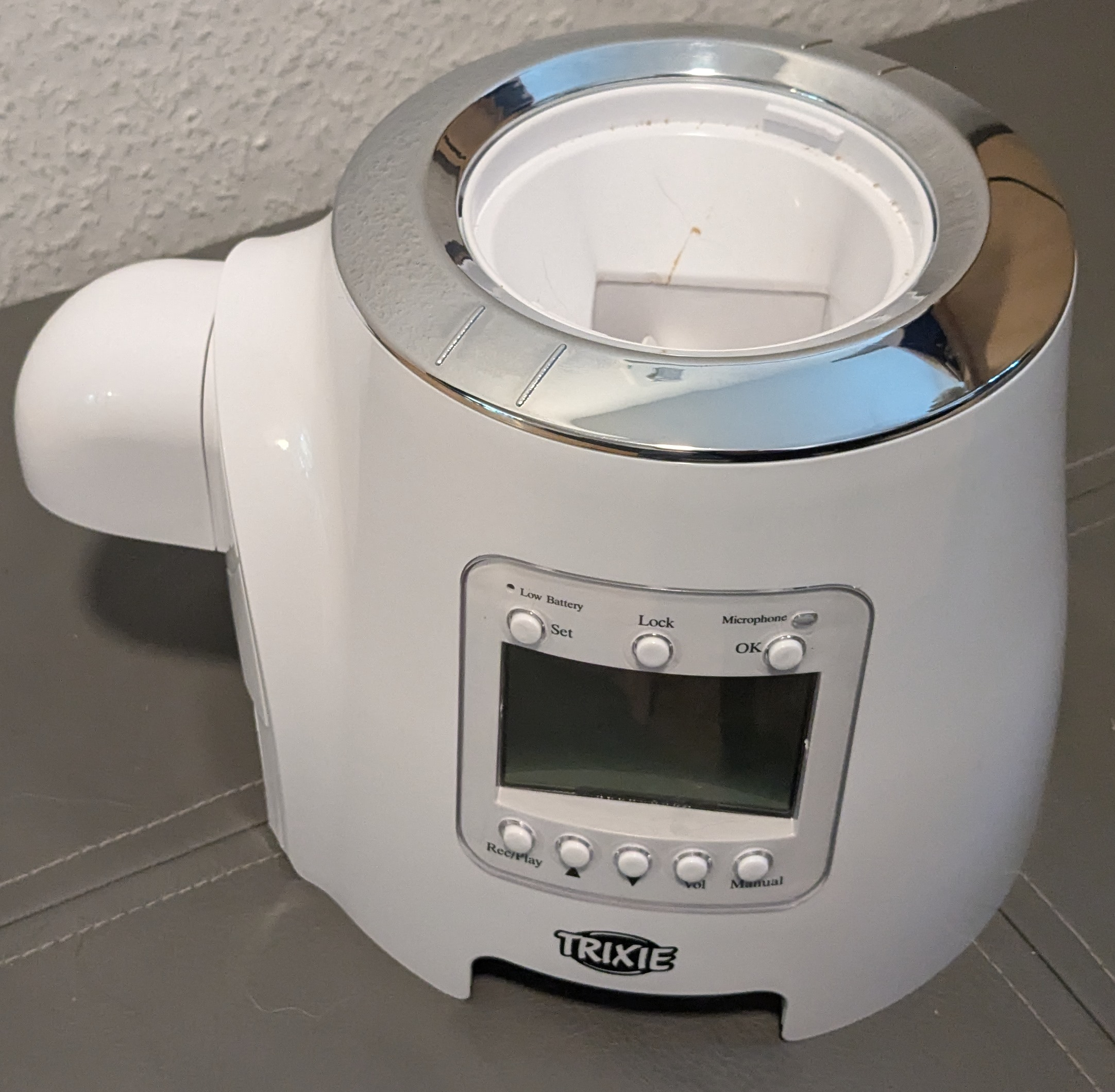 Just the core of the feeder is shown. It has a silver ring where it would normally meet the hopper and an inlet for food can be seen on the top. On the side facing us, we can see a blank LCD screen with three buttons above and five below.The top buttons from left to right are labeled “set”, “Lock”, and “OK”. The bottom buttons from left to right are: “Rec/Play”, an arrow pointing upwards, an arrow pointing downwards, “Vol” and “Manual”.