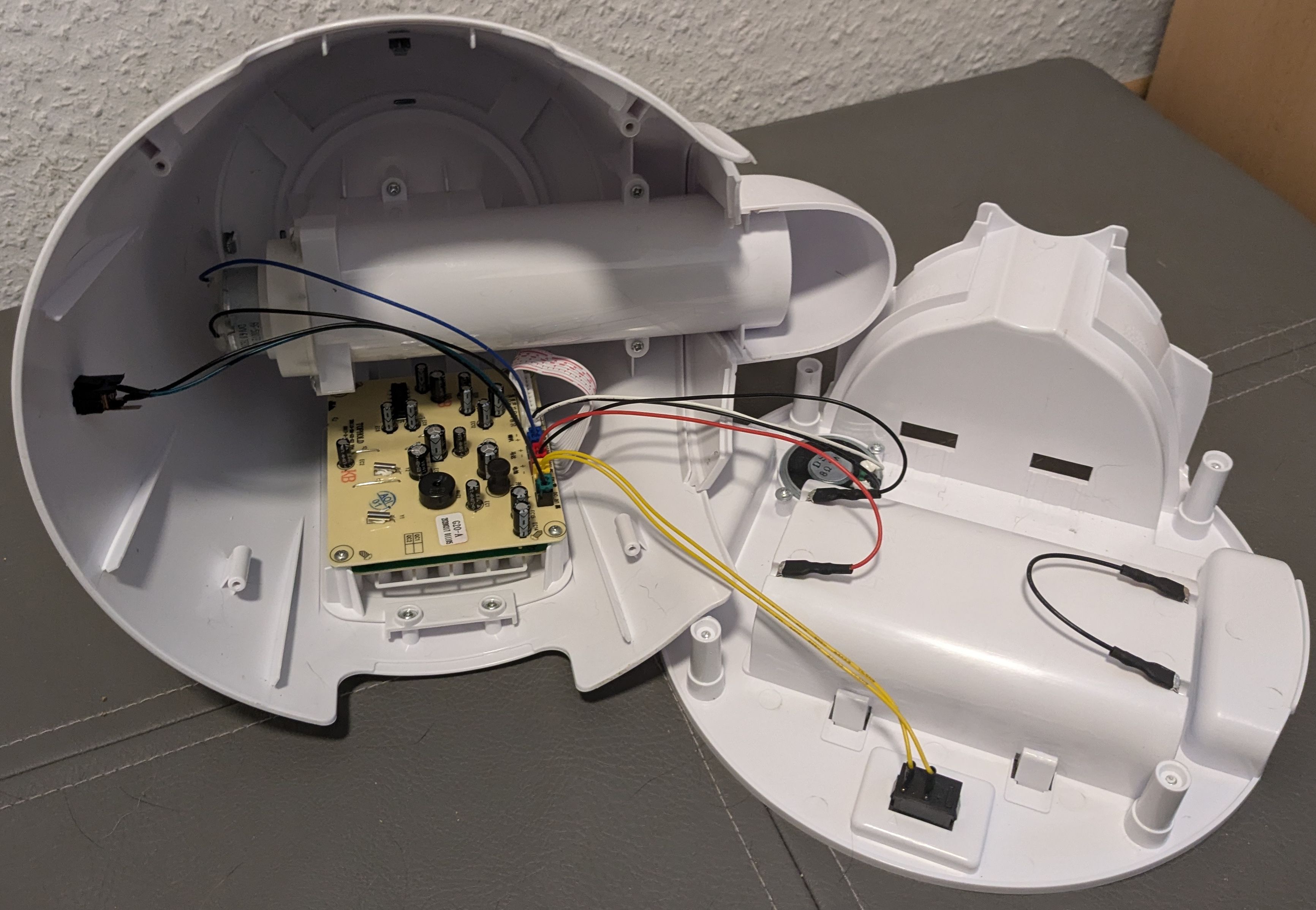 The inside of the feeder is shown. The core is in two pieces, with the bottom on the right-hand side and the top on the left. Inside the top, a tube can be seen leading to the spout. Additionally, we can see a circuit board with wires leading to the back of the tube, a point on the outside of the top and to the base. On the base we can see that yellow wires lead to a black box, that a black and red pair leads to a raised area on the base, and that a white and black pair leads to the back of a speaker.