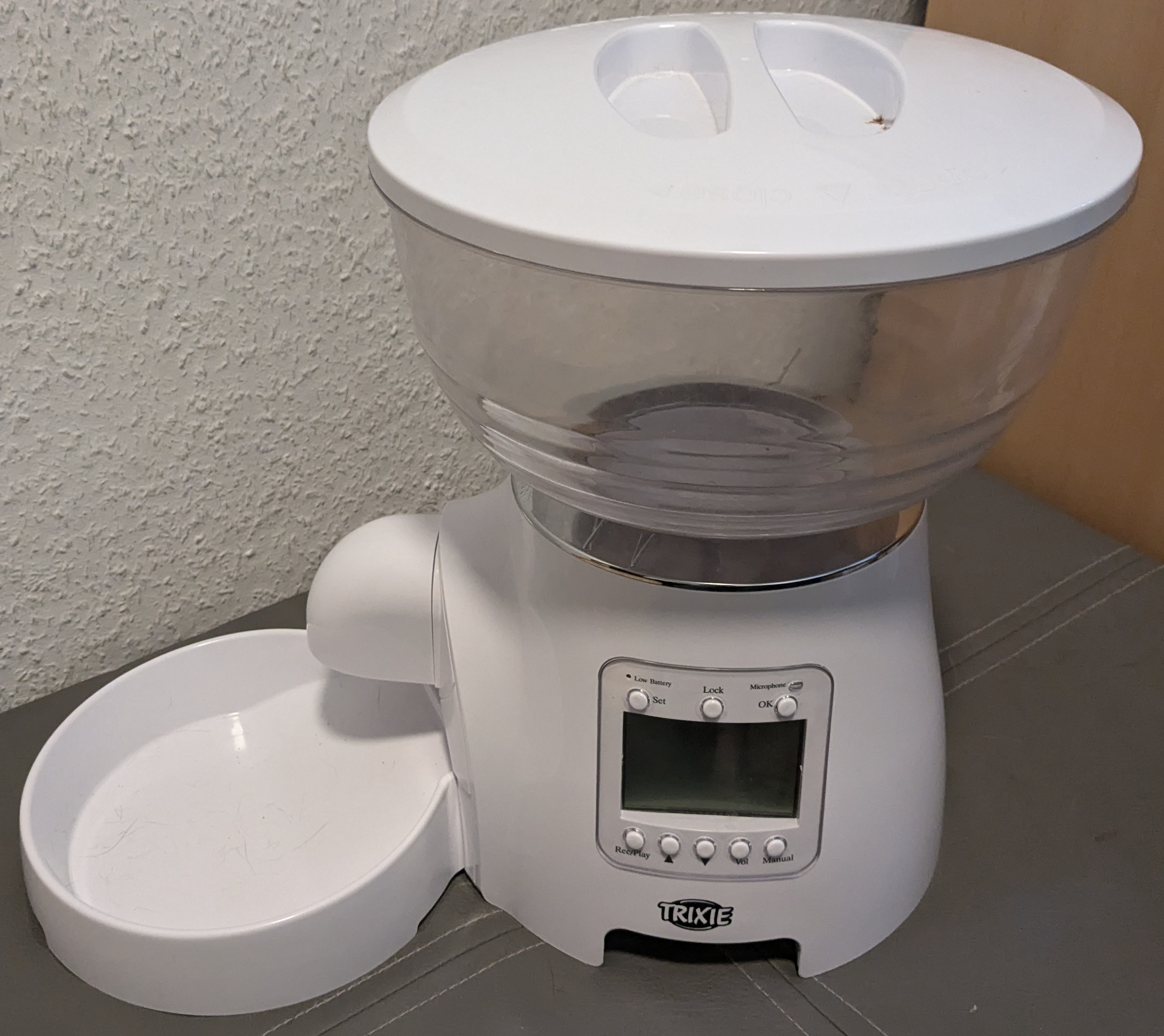 A fully assembled cat feeder. It consists of a flat, white, plastic bowl, attached to the front of an upright white, plastic dome. The upright dome (referred to as the core) has an LCD screen and buttons (described in more detail in later alt texts) and a spout that comes out over the bowl. On top of the upright dome is an upside-down clear plastic dome with a white top - the hopper.